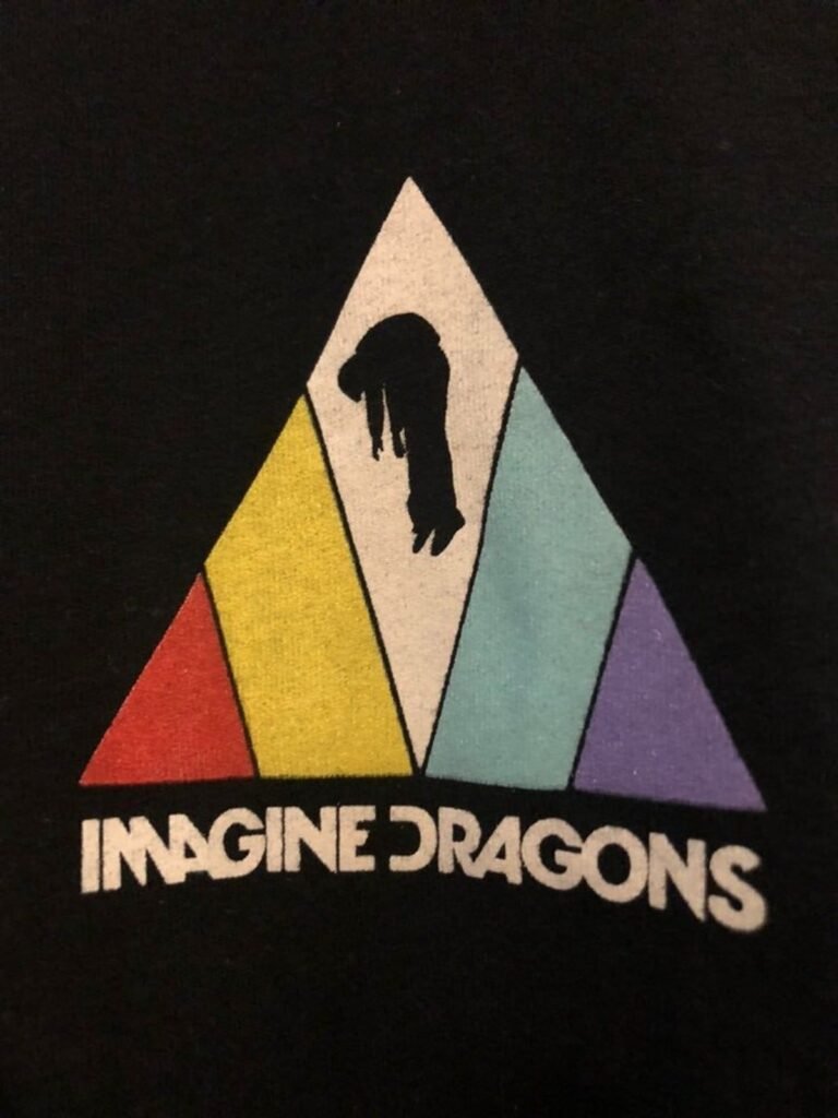 An Imagine Dragons Evolve Era Logo featured on a black background. The design showcases a large triangle divided into colorful vertical sections of red, yellow, white, light blue, and purple. Inside the central white section, a black silhouette of a person is shown suspended in mid-air. Below the triangle, the "Imagine Dragons" band name is printed in its signature bold, white sans-serif typography.